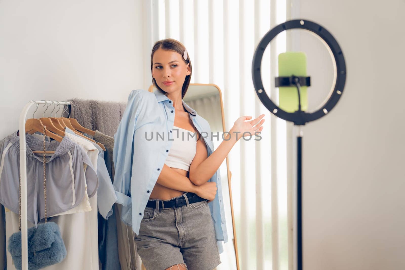 Young woman making fashion video content for social media. Blogger smiles to camera using selfie stick and light ring while making persuasive online clothing sell to audience or followers. Unveiling