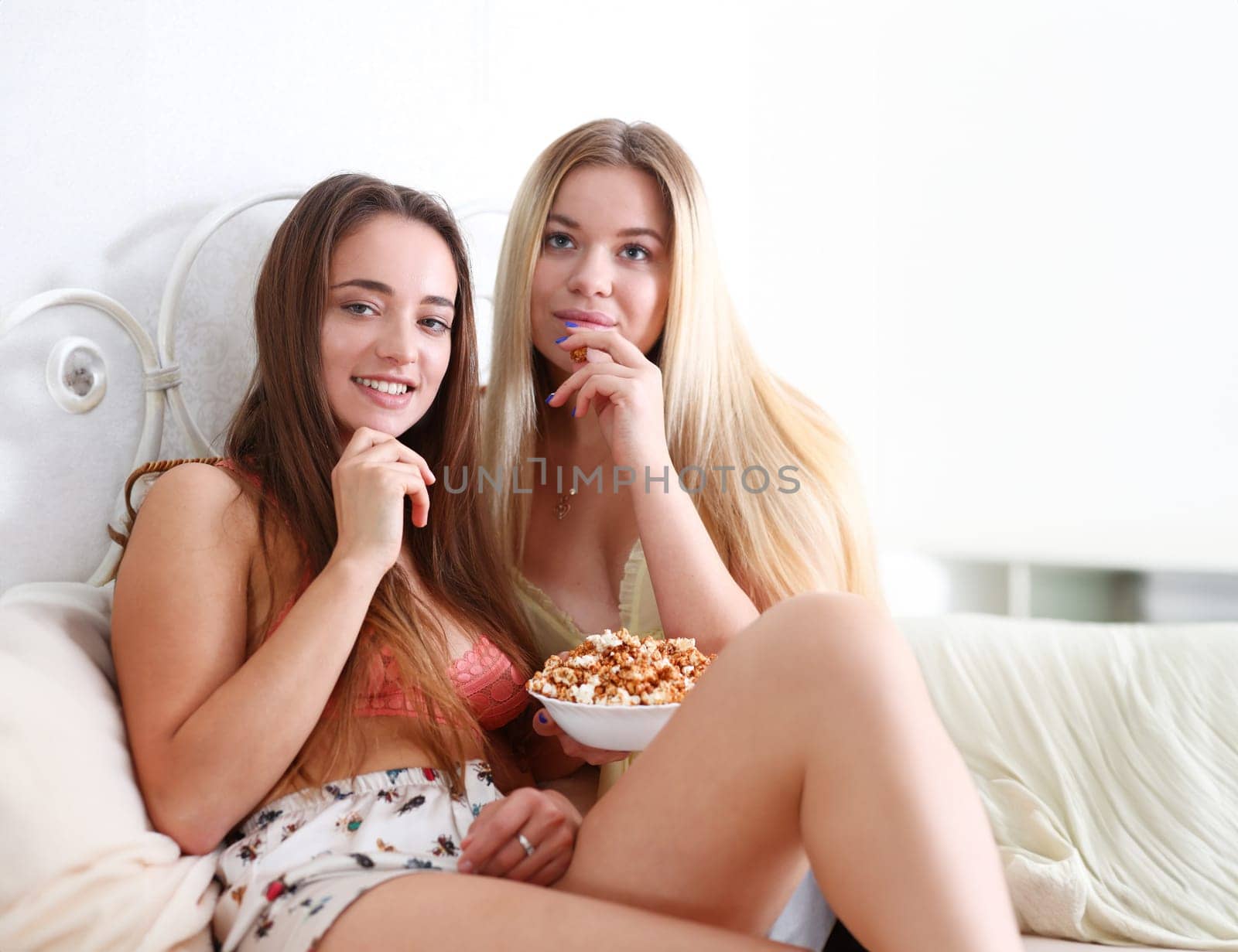 Portrait of two beautiful young smiling woman friends watching a comedy movie melodramma in an apartment eating popcorn and laughing on the bed in pajamas during a hen party switch channels tv.