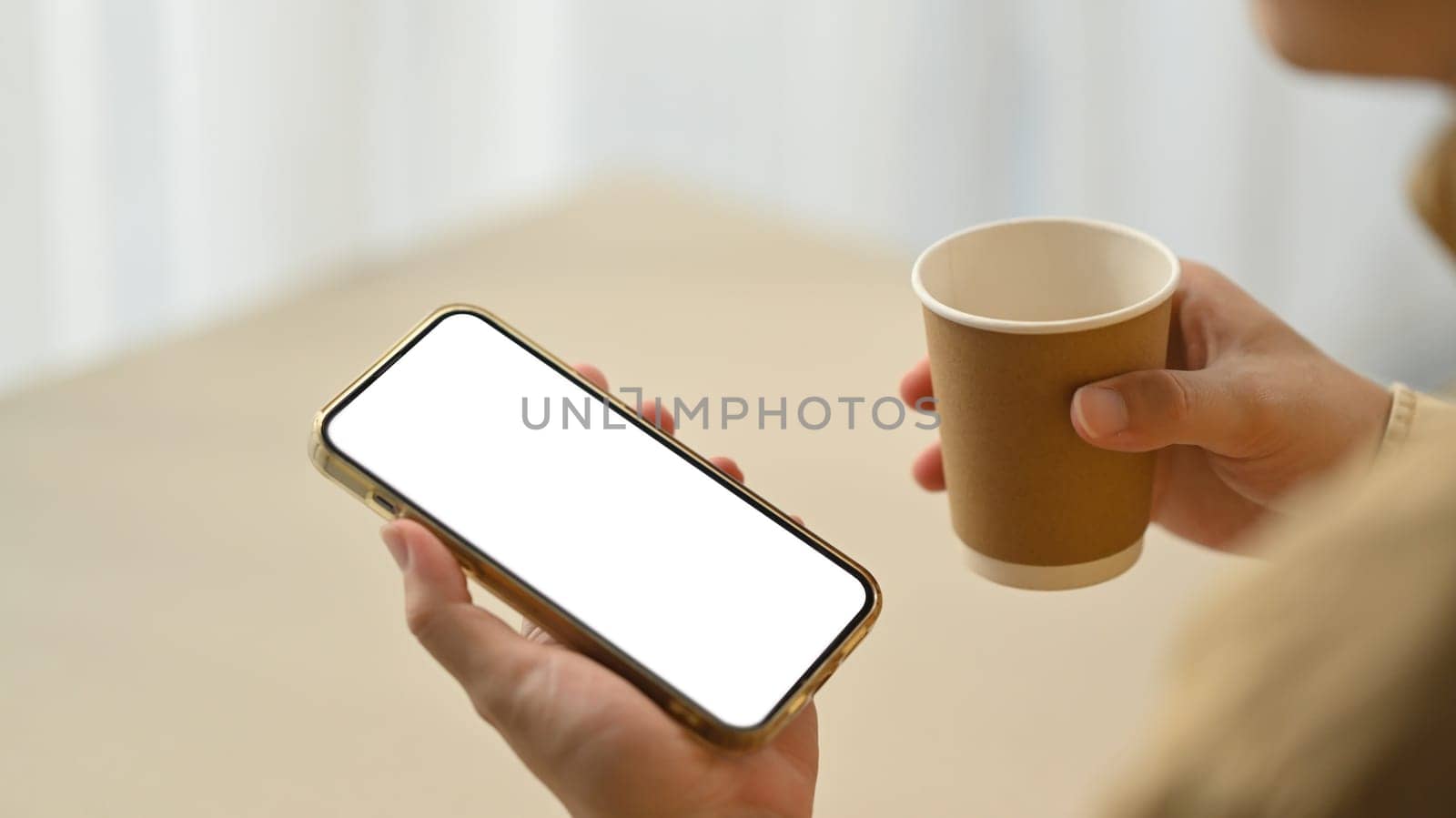 Closeup woman using mobile phone and drinking coffee in bright office. Empty screen for advertising.