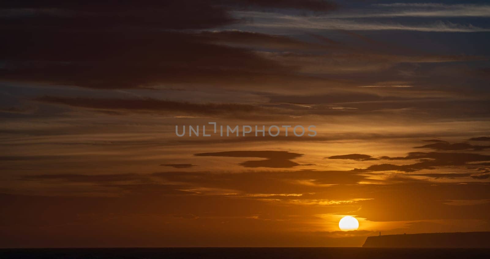 Orange sunset creates a scenic silhouette of a lighthouse on a cliff.