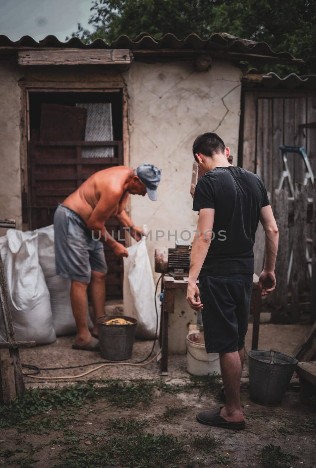 Two Caucasian men, old and young, are manually making dry compound feed for pets in a homemade crusher, standing on the street of a village house in the countryside, close-up side view in dark style and selective focus.