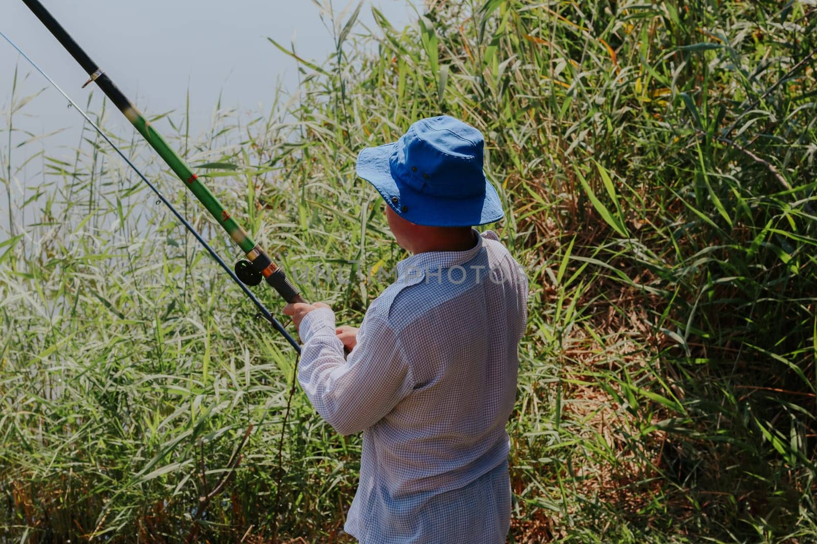 One Caucasian young man in old dirty clothes with a blue hat on his head catches fish with a fishing rod standing on the shore of a lake in the tall grass on a sunny summer day, close-up side view.