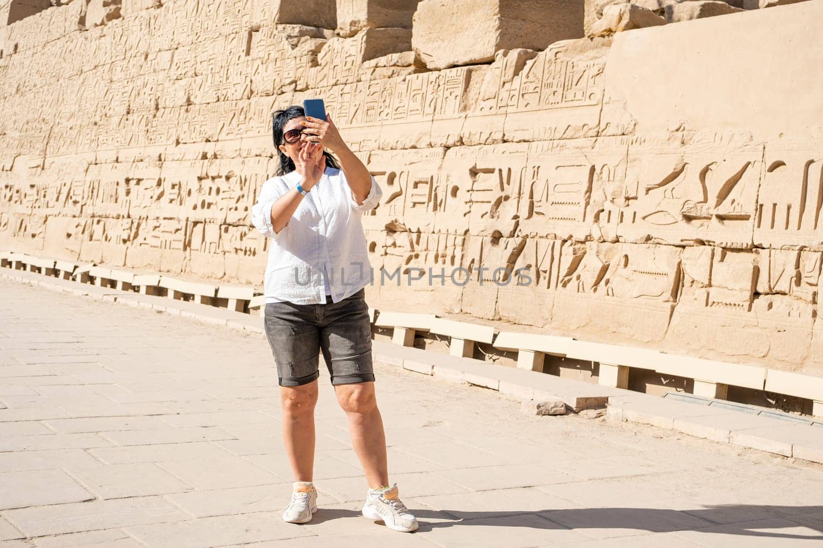 mature adult woman traveler explores the ruins of the ancient Karnak temple in the city of Luxor in Egypt, taking photo using smartphone. Great row of columns with carved hieroglyph