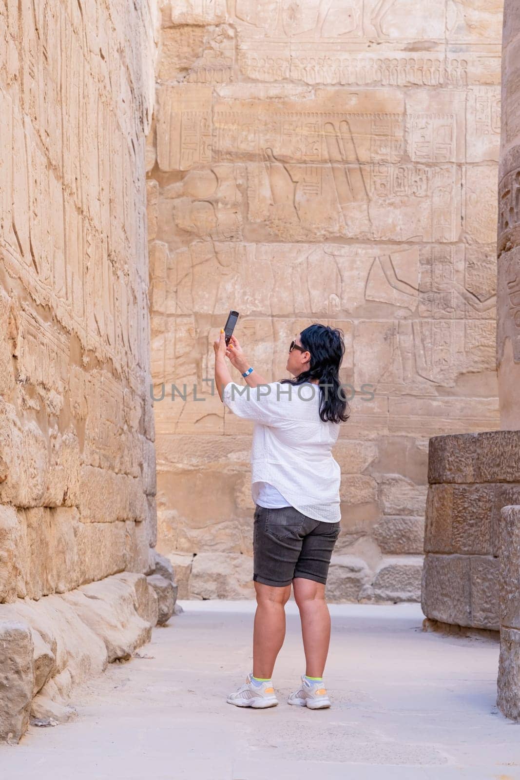 mature adult woman traveler explores the ruins of the ancient Karnak temple in the city of Luxor in Egypt, taking photo using smartphone. Great row of columns with carved hieroglyph
