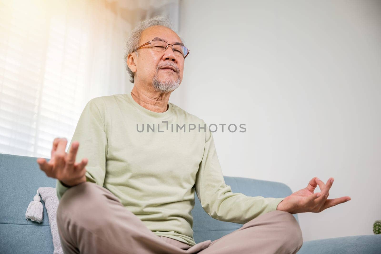 senior man sitting on sofa in living room holding hands in mudra practicing home yoga in lotus pose by Sorapop