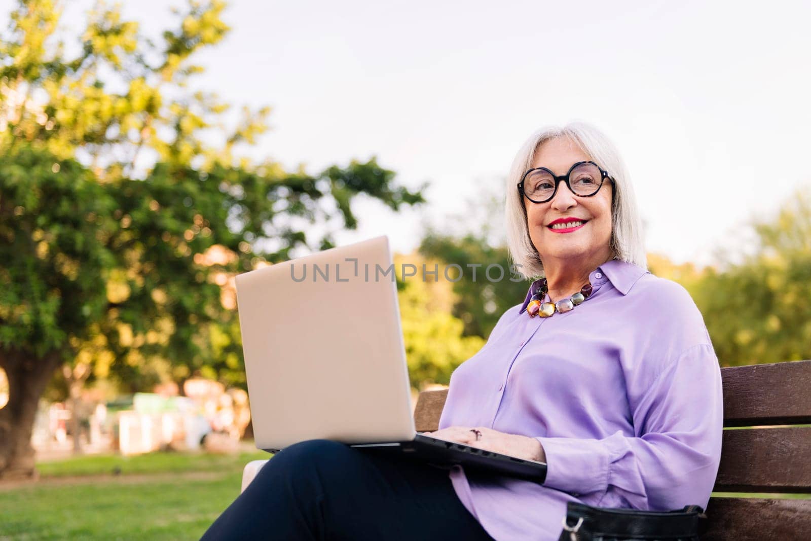 smiling senior woman using laptop outdoors sitting on park bench, concept of technology and elderly people leisure, copy space for text