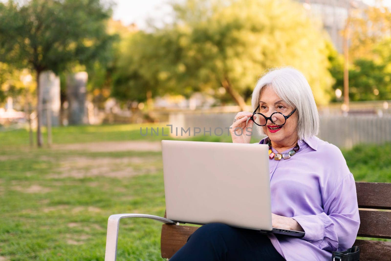 beautiful senior woman with glasses using laptop outdoors sitting on park bench, concept of technology and elderly people leisure, copy space for text