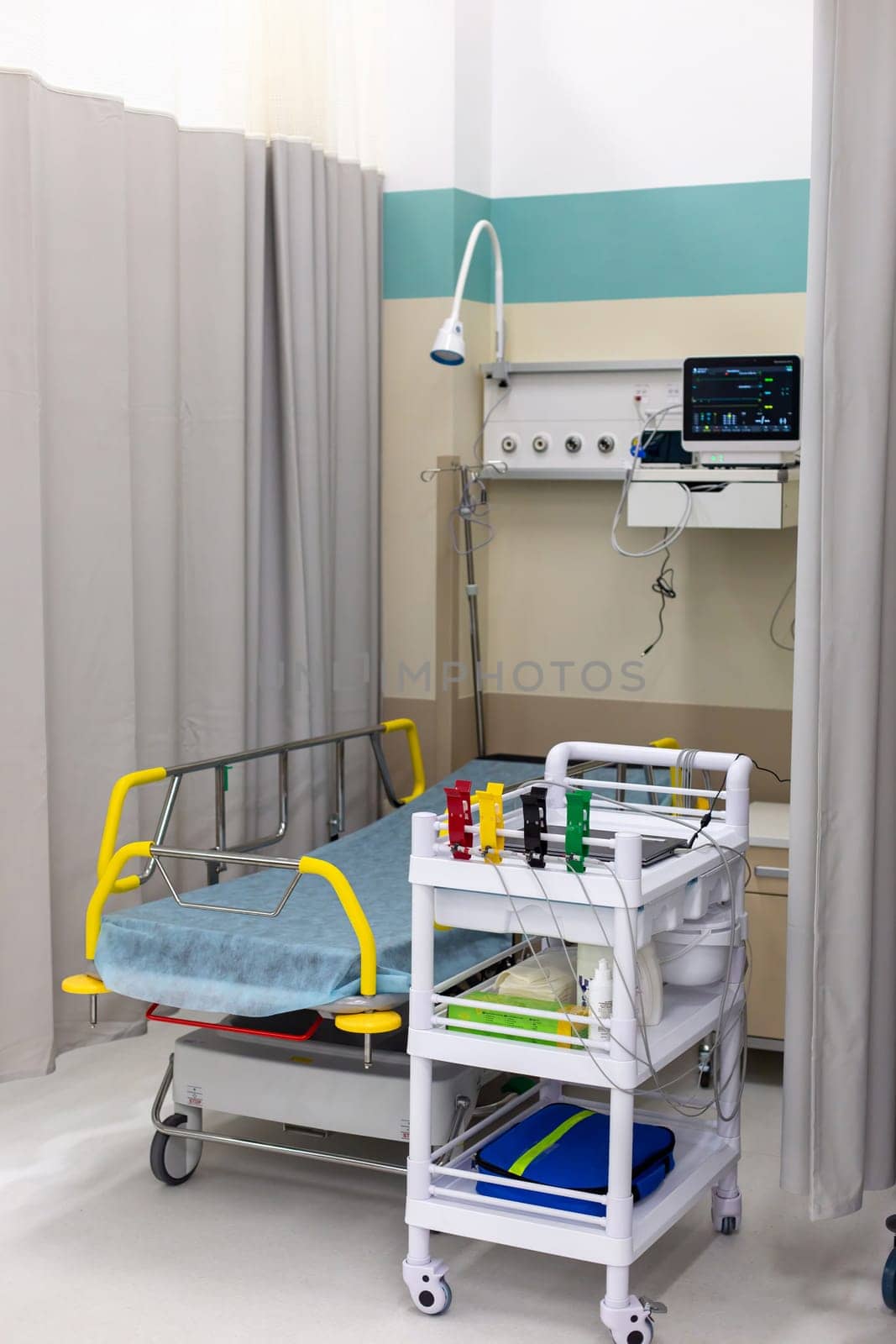 Hospital room setup with an empty patient bed, a medical trolley, and a vital signs monitor against a curtained backdrop