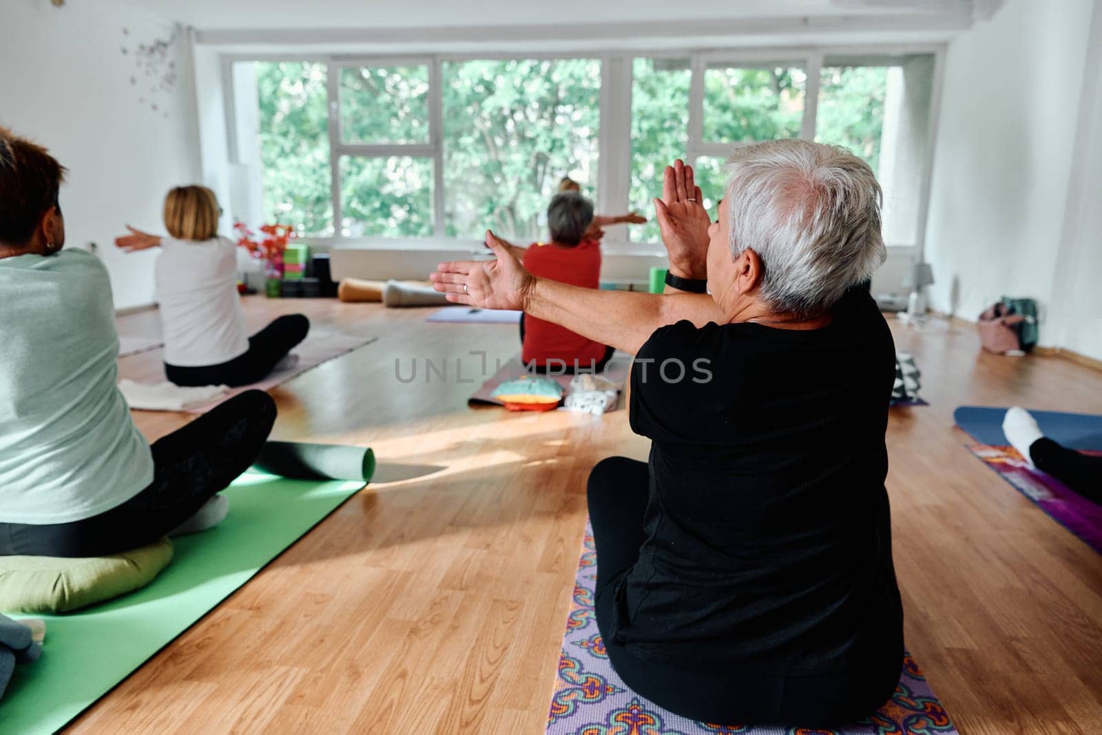 A group of senior women engage in various yoga exercises, including neck, back, and leg stretches, under the guidance of a trainer in a sunlit space, promoting well-being and harmony by dotshock