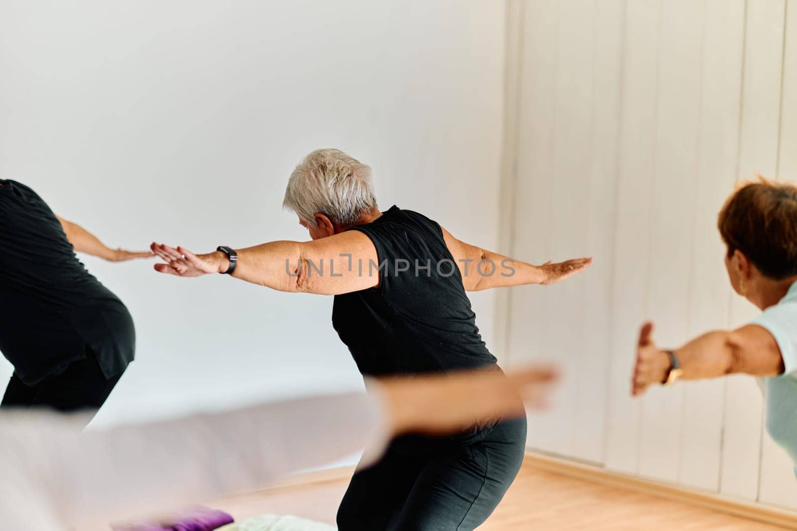 A group of senior women engage in various yoga exercises, including neck, back, and leg stretches, under the guidance of a trainer in a sunlit space, promoting well-being and harmony.