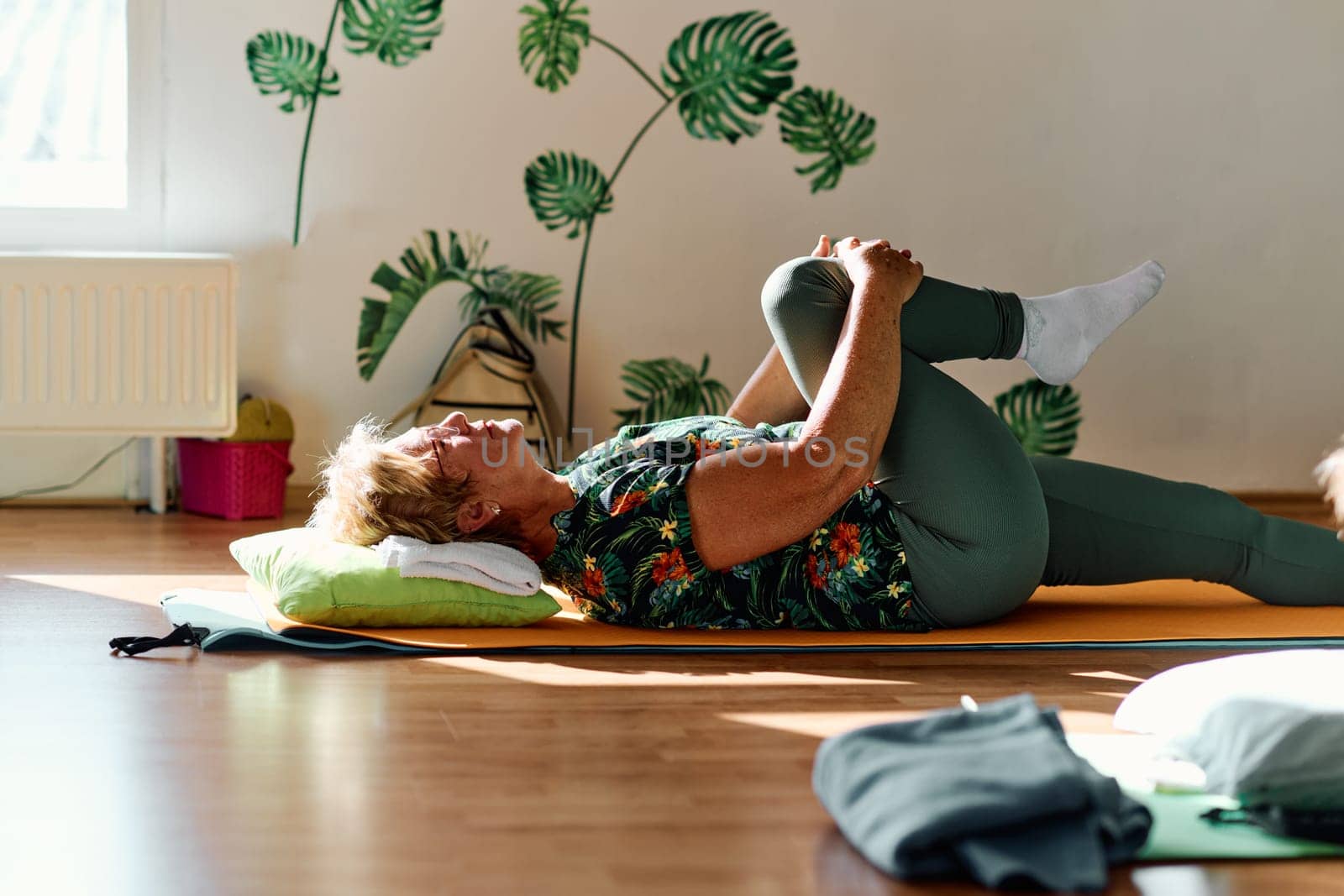 In a sunlit space, a senior woman gracefully practices rejuvenating yoga, focusing on neck, back, and leg stretches, embodying serenity and well-being by dotshock