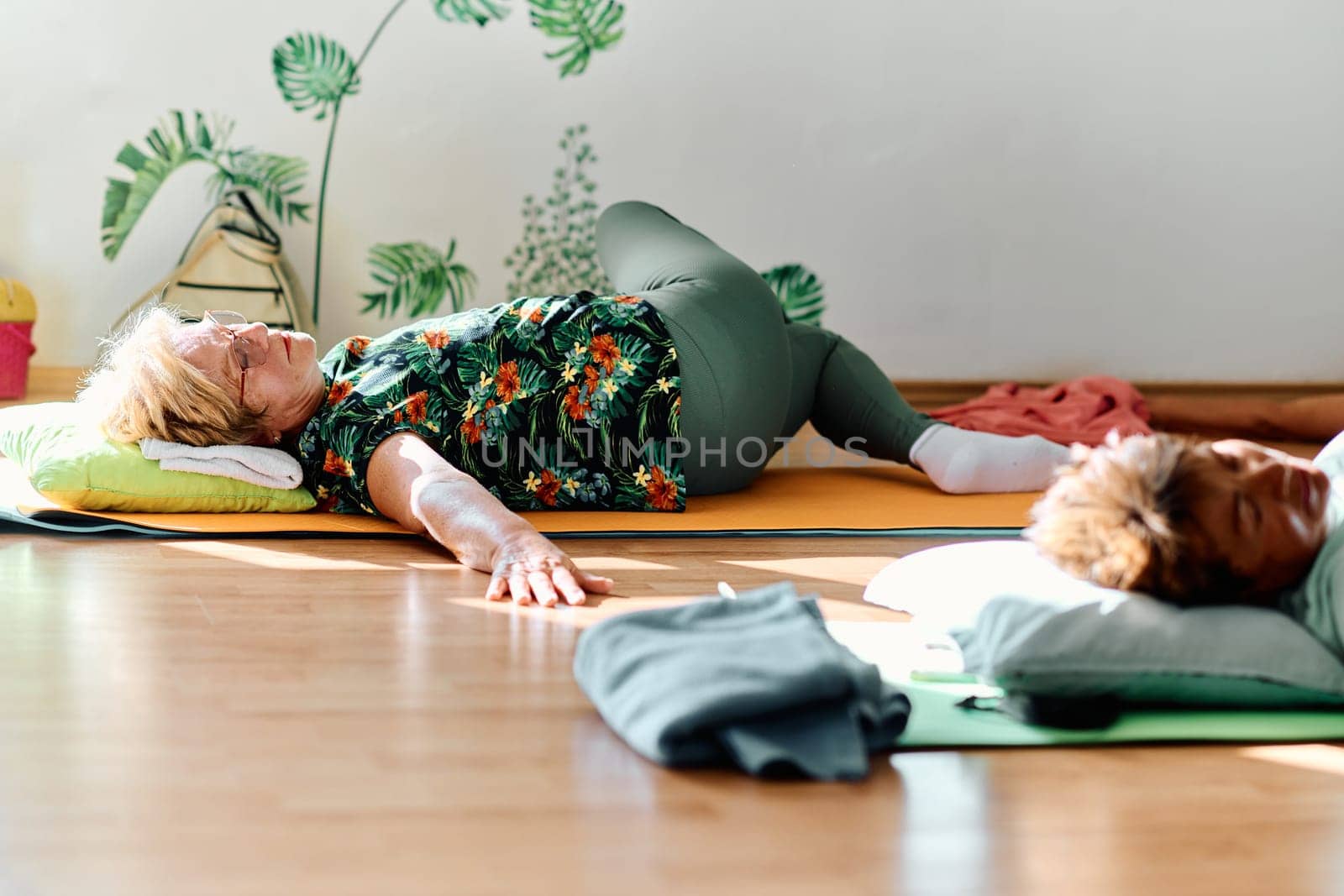 A group of senior women engage in various yoga exercises, including neck, back, and leg stretches, under the guidance of a trainer in a sunlit space, promoting well-being and harmony by dotshock