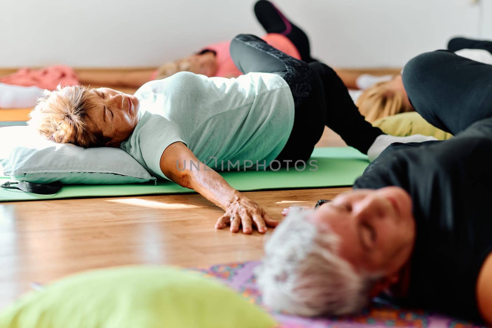 A group of senior women engage in various yoga exercises, including neck, back, and leg stretches, under the guidance of a trainer in a sunlit space, promoting well-being and harmony by dotshock