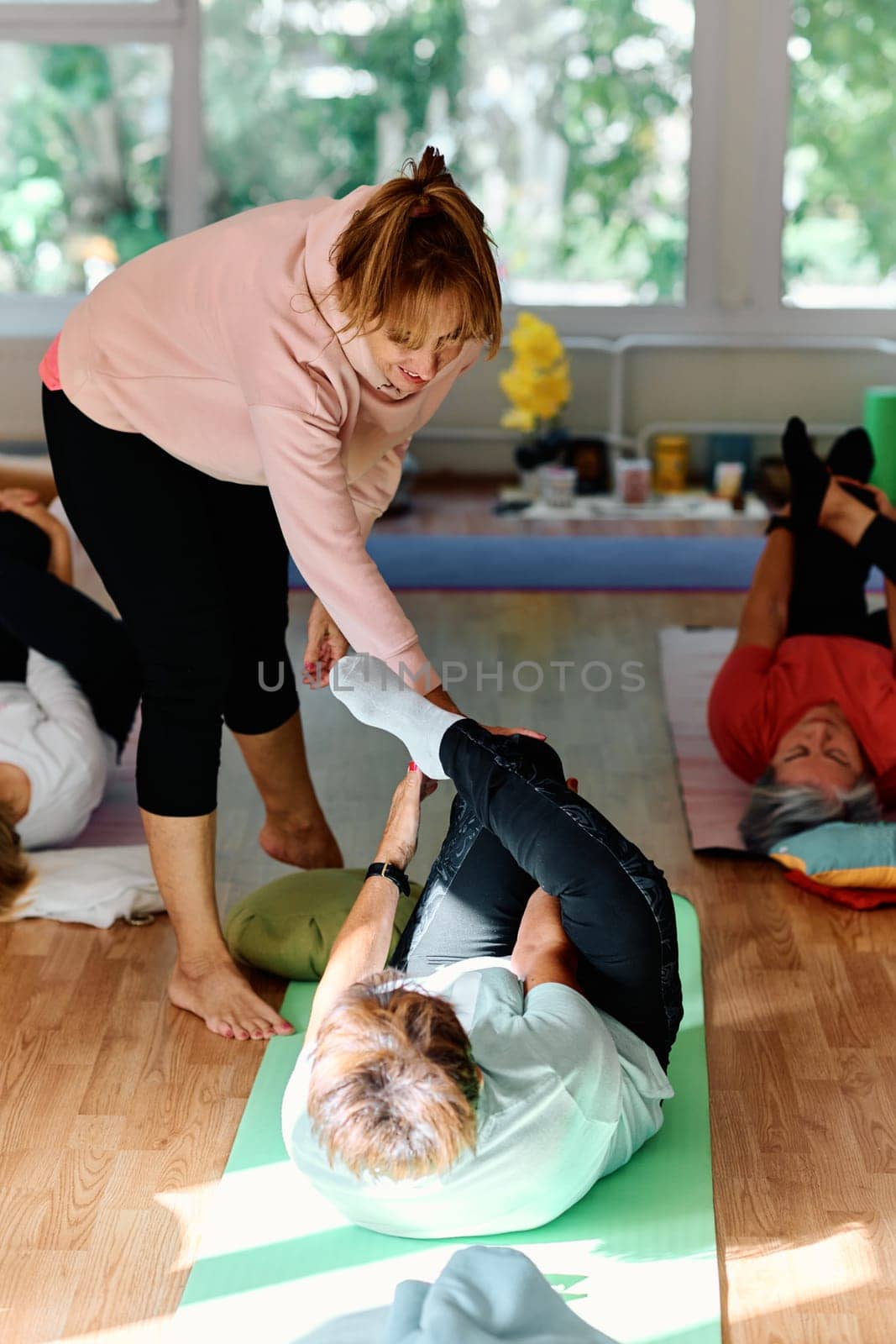 A skilled trainer oversees a group of senior women practicing various yoga exercises, including neck, back, and leg stretches, in a sunlit space, promoting wellness and harmony.