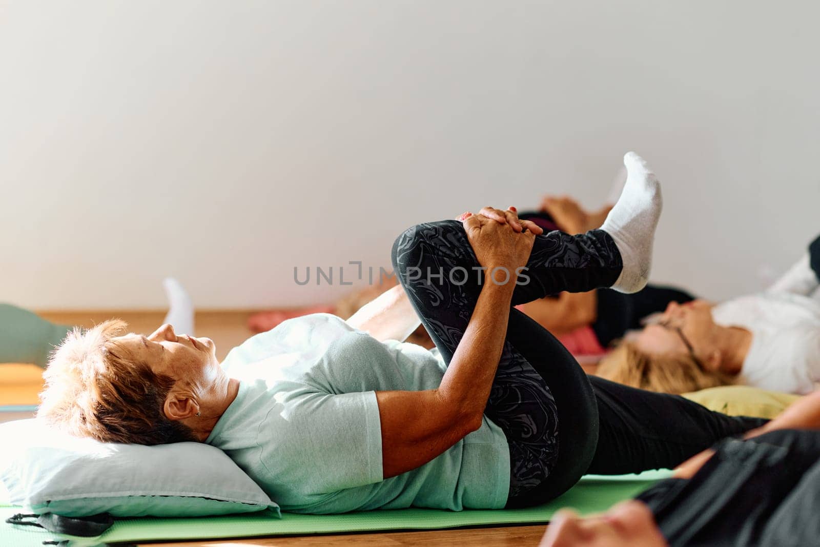 In a sunlit space, a senior woman gracefully practices rejuvenating yoga, focusing on neck, back, and leg stretches, embodying serenity and well-being by dotshock