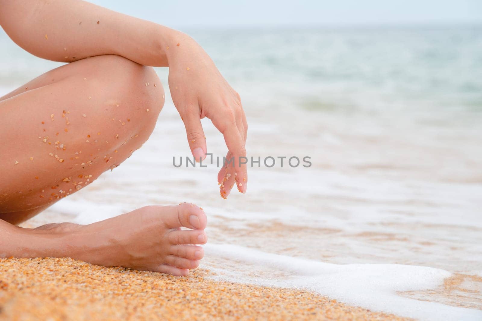 A woman sits on the shore and relaxes. A woman's leg and hand on her knee, a girl sitting on the sea sand on the beach on a summer day, close-up