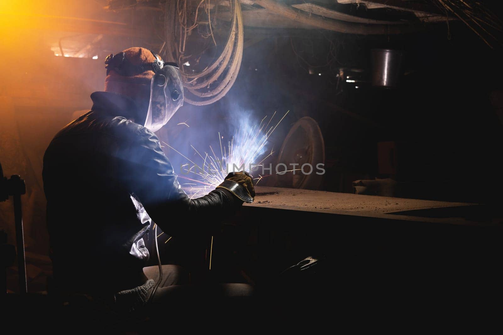 A welder in a workshop welds a metal part. General plan of an old cluttered garage where a man in protective gloves and a mask is making.