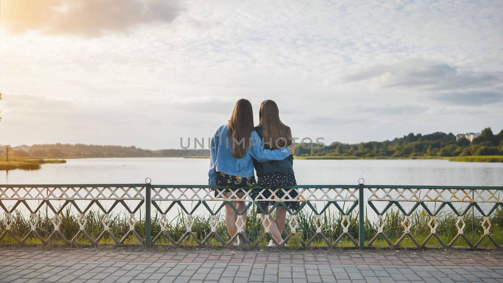 Two girlfriends sitting in an embrace on the lake shore. by DovidPro