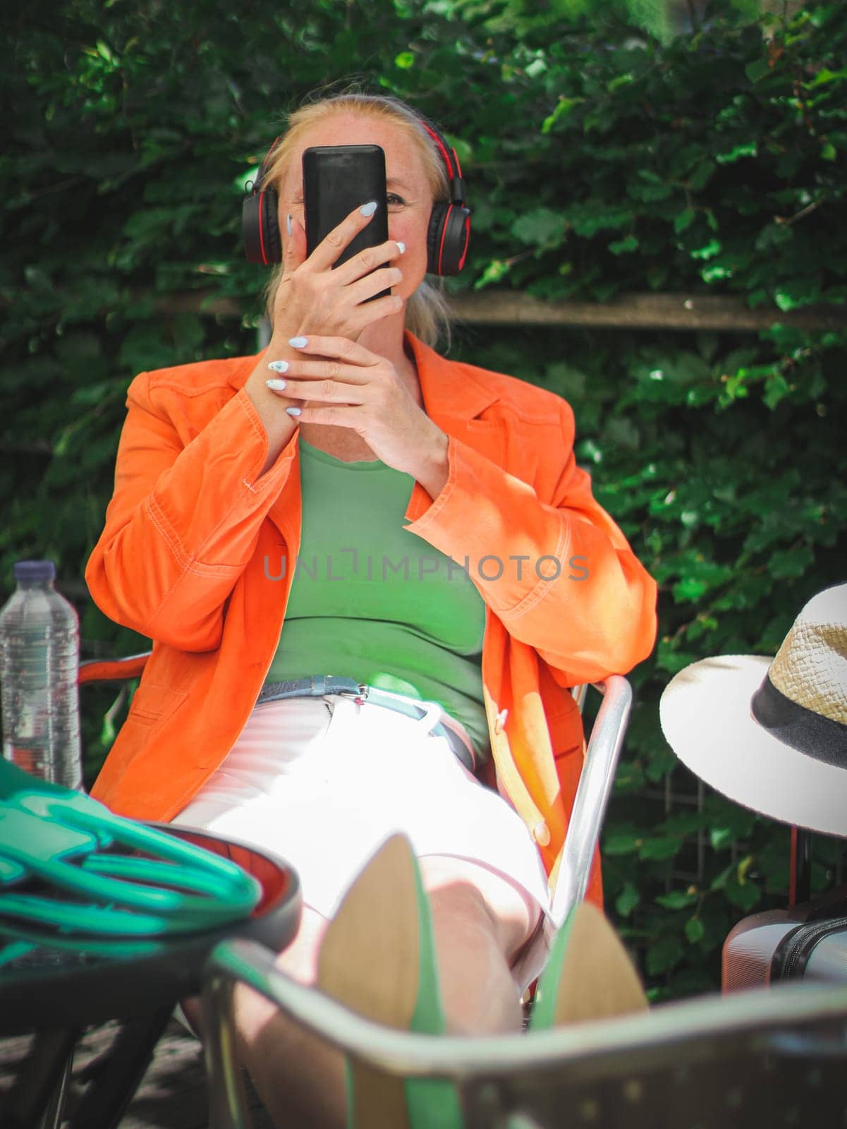 A young beautiful stylish Caucasian blonde woman in a bright orange jacket with a green bag, headphones and a suitcase holds a smartphone in front of her face, having fun communicating, sits with her legs stretched out on a chair in a street cafe, close-up side view. Technology use concept.