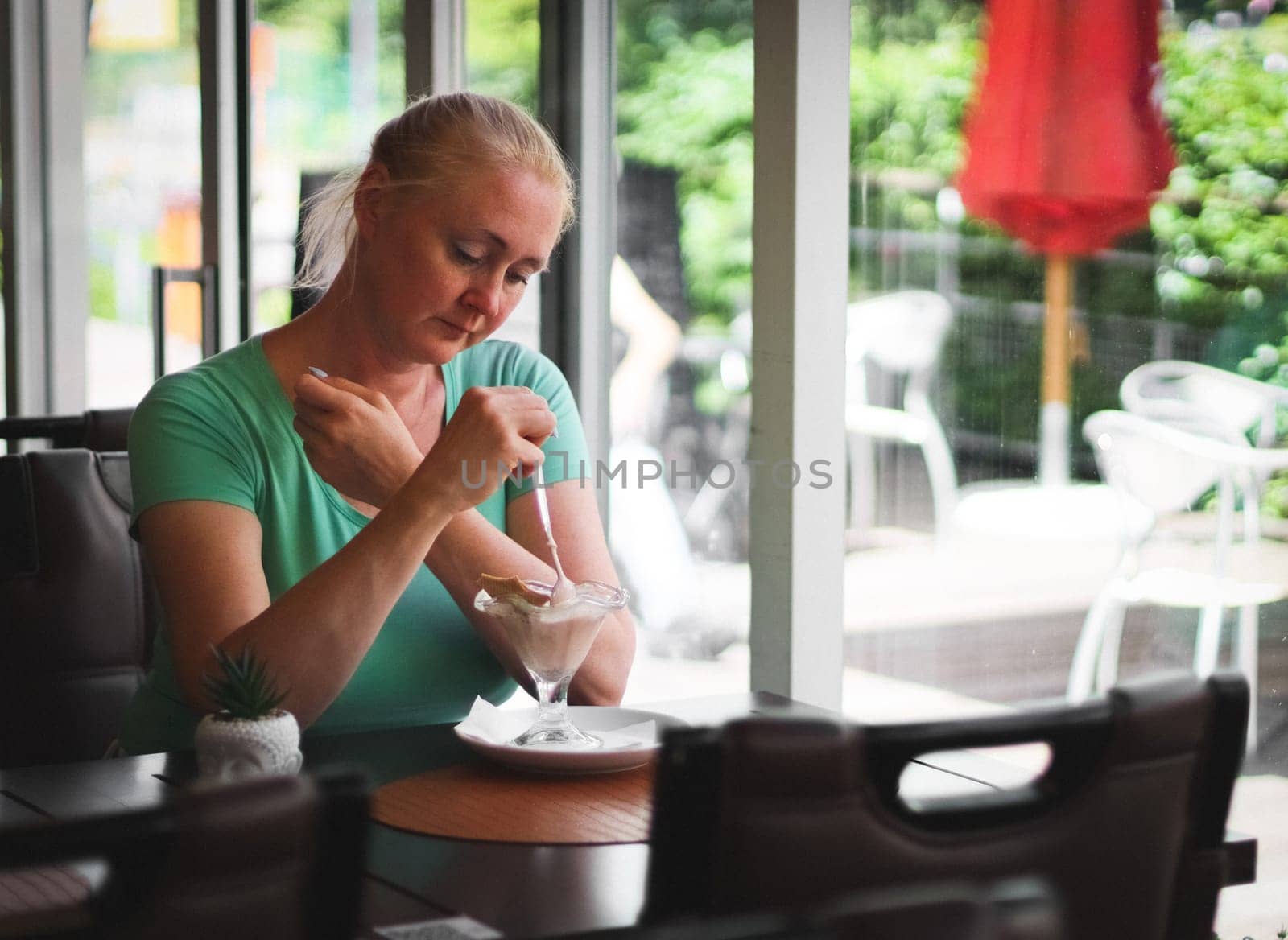 One young Caucasian very sad woman sits at a table in a cafe and eats a dessert spoon of melted strawberry ice cream with a piece of waffle, close-up side view. Concept of eating ice cream, summer cold dessert.