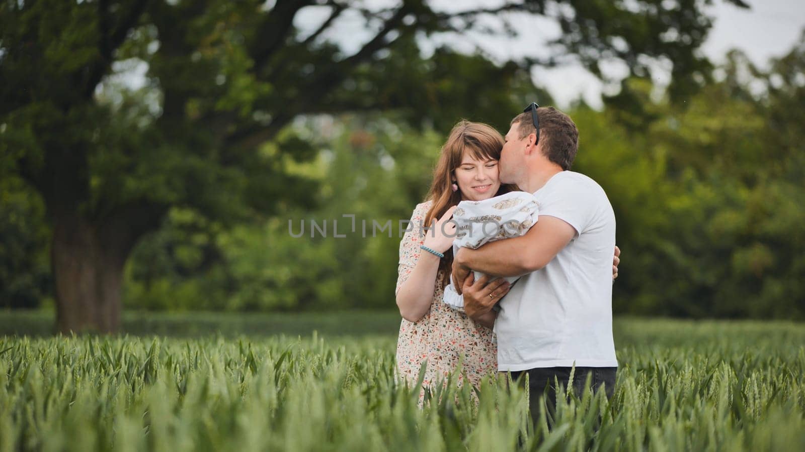 A young couple standing in a field kissing and holding their newborn baby in their arms