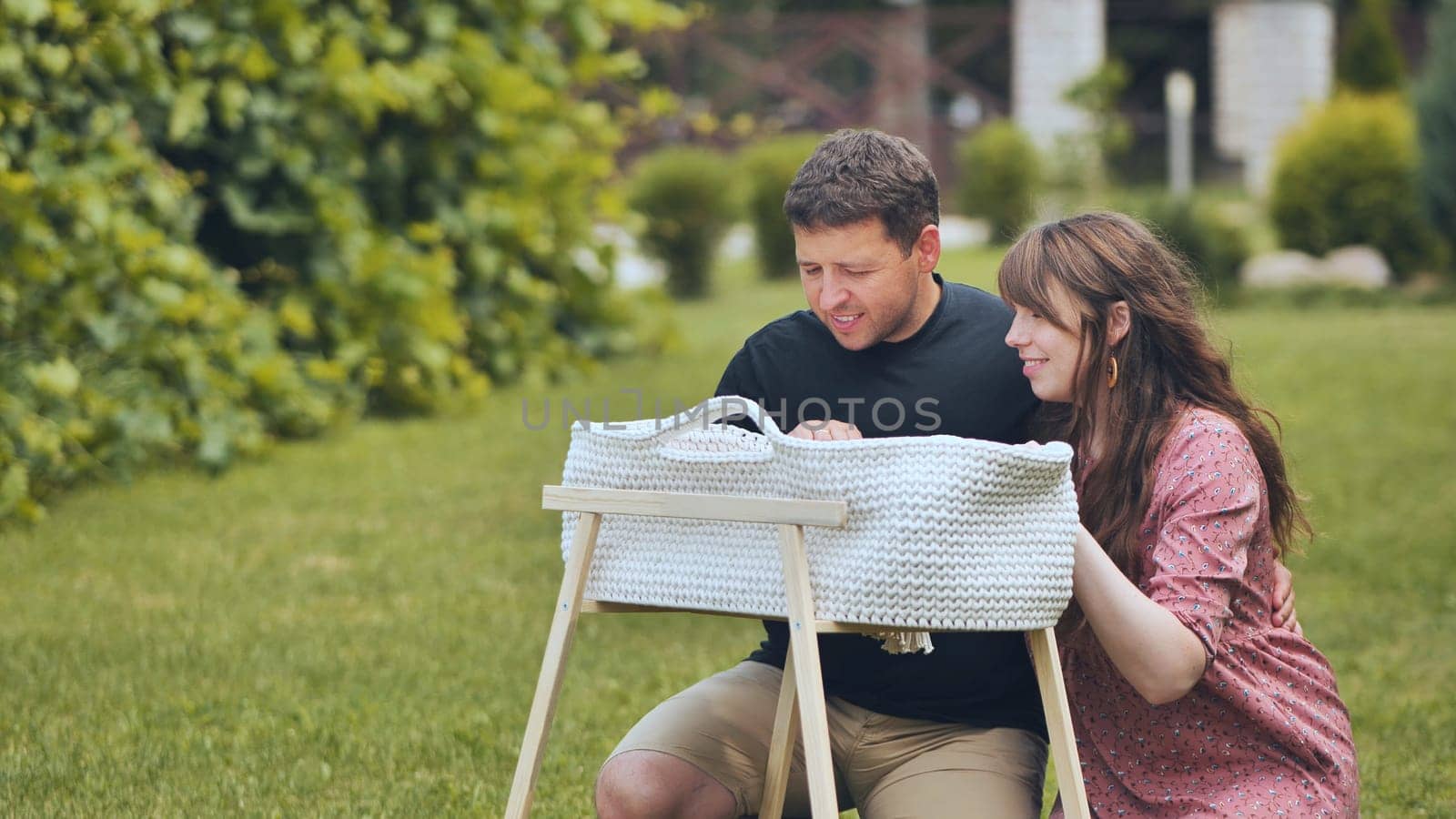 A young couple looks at their newborn baby in a cradle in the garden. by DovidPro