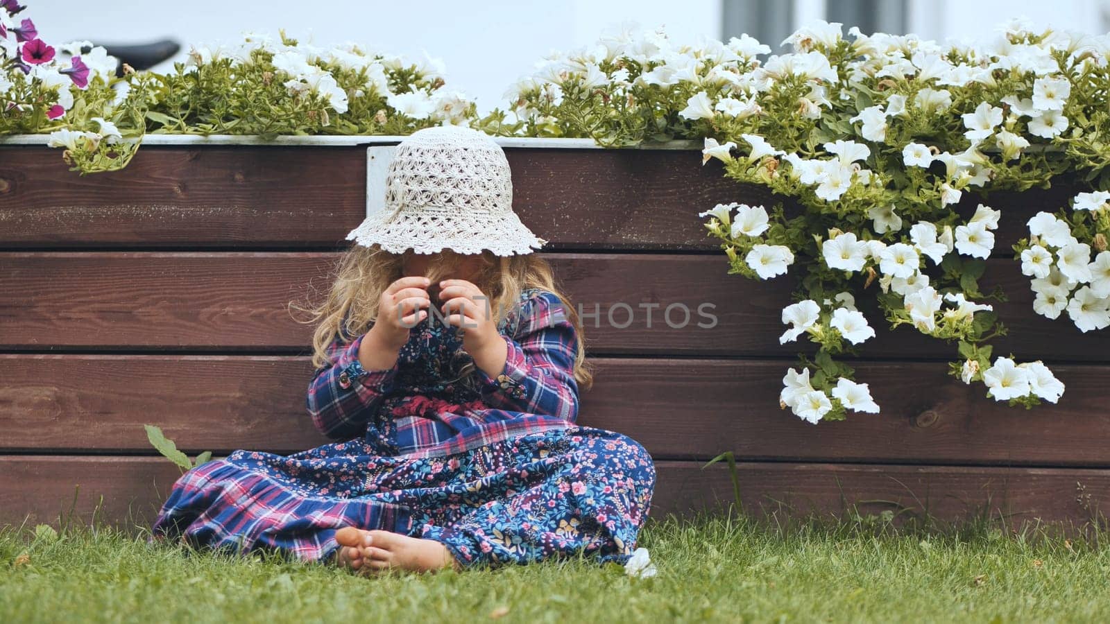 A little girl in a panama hat sits by the white petunias in the garden