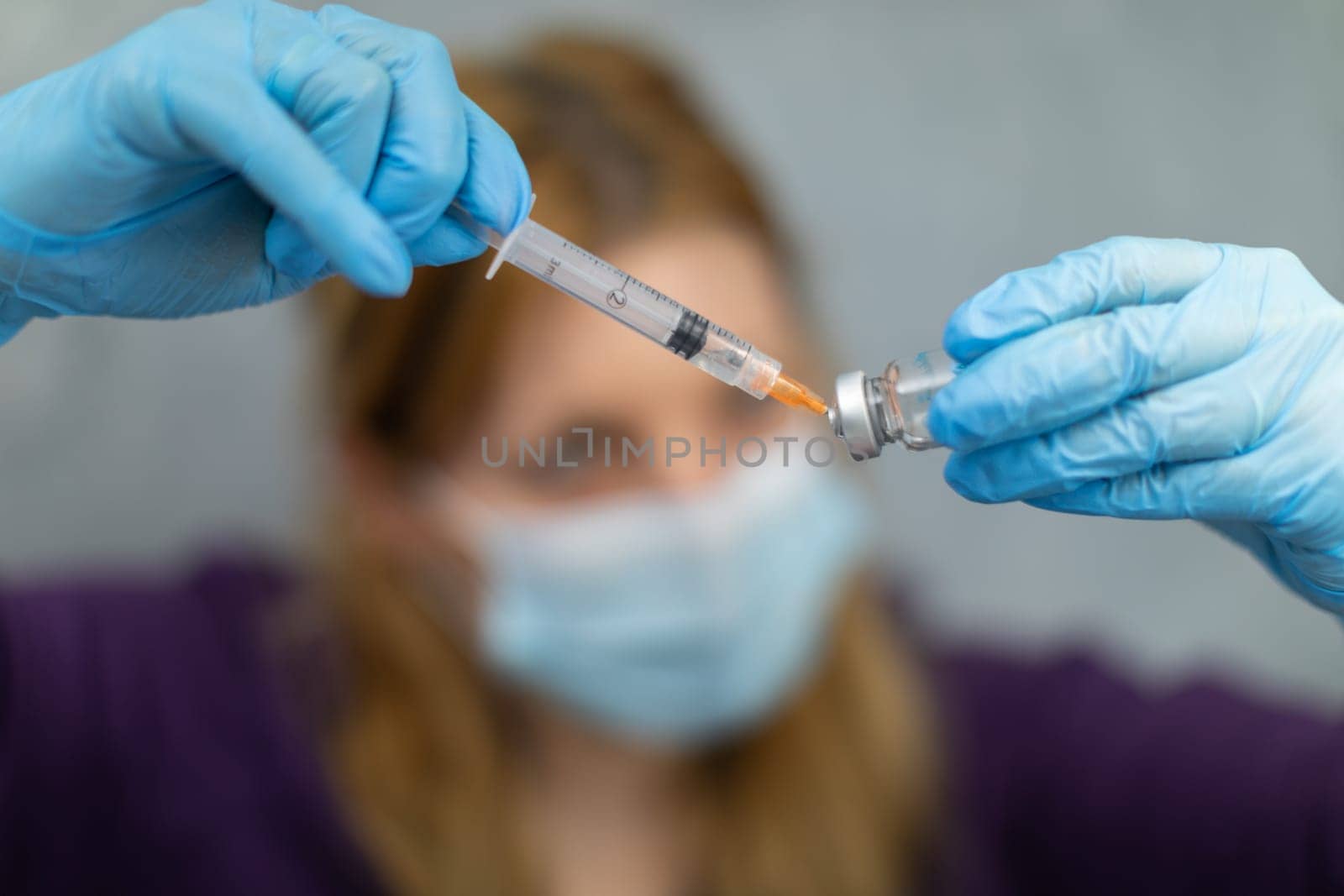 Blurred foreground. The sharpened foreground shows hands wearing disposable gloves. One hand is holding a vial of medicine, while the other is acquiring the drug using a syringe with a needle.