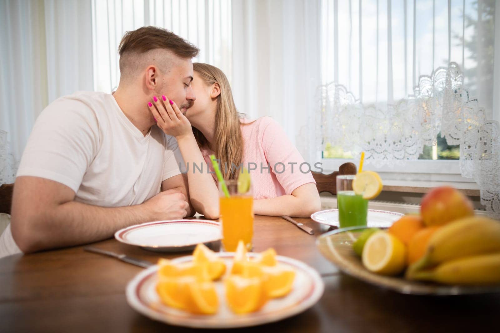 A girl kisses her boyfriend on the cheek. The couple sits at a table against the backdrop of a window. Young people meeting on a date. They are sitting at a table with fruit on it.