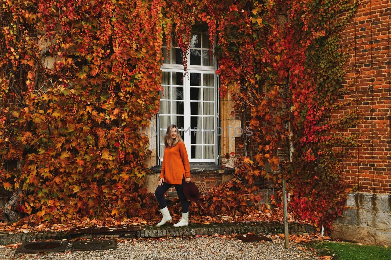 Woman in an orange sweater and a hat near a wall with wild grapes in autumn by Godi