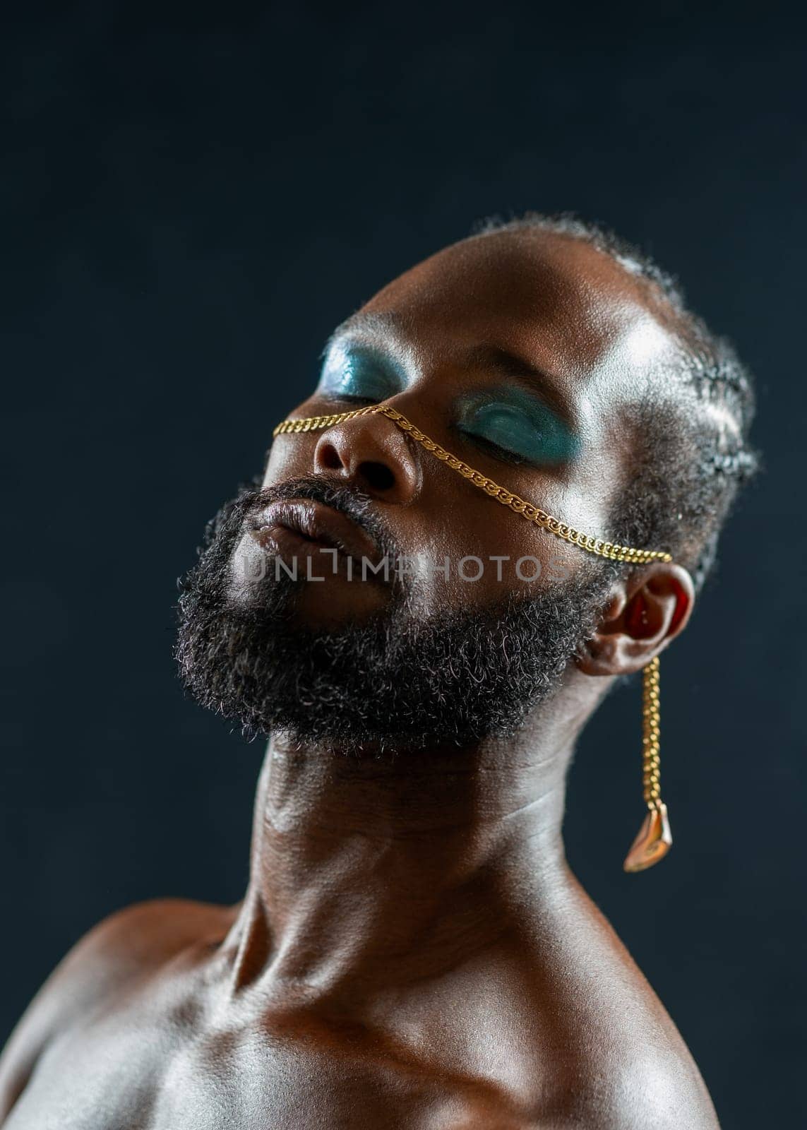 Portrait of shirtless African American gay man with blue eyeshadows and golden chain on face standing eyes closed against dark blue background