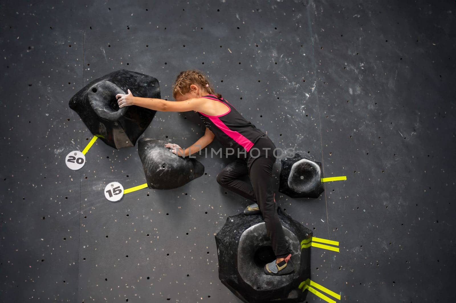 Side view of determined girl climbing artificial wall. Brave and strong female child climbs up mock wall. Full body of sporty girl at bouldering center.