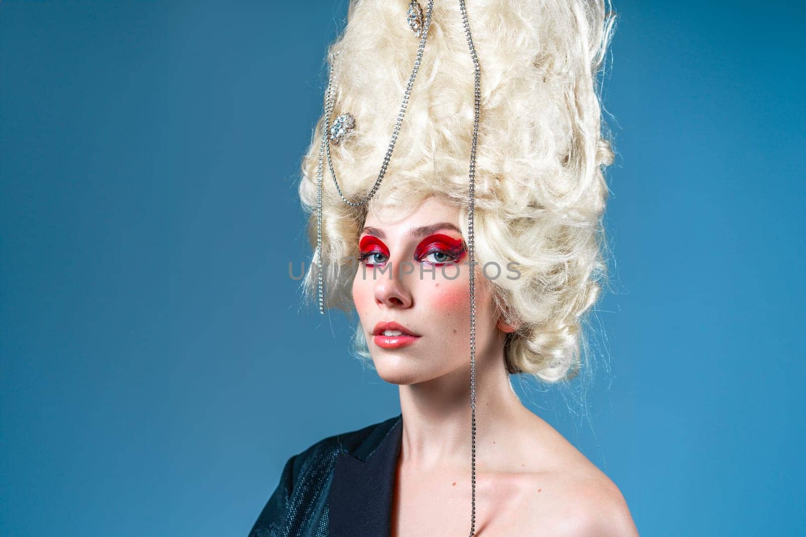 Portrait of beautiful young female model with vintage royal wig and red makeup looking at camera over blue background