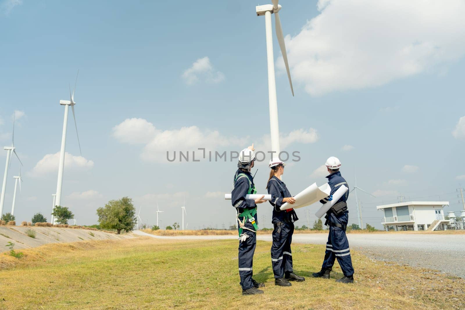 Group of professional technician workers stand with holding project paper plan in front of wind turbine or windmill and look back in area for power plant business.