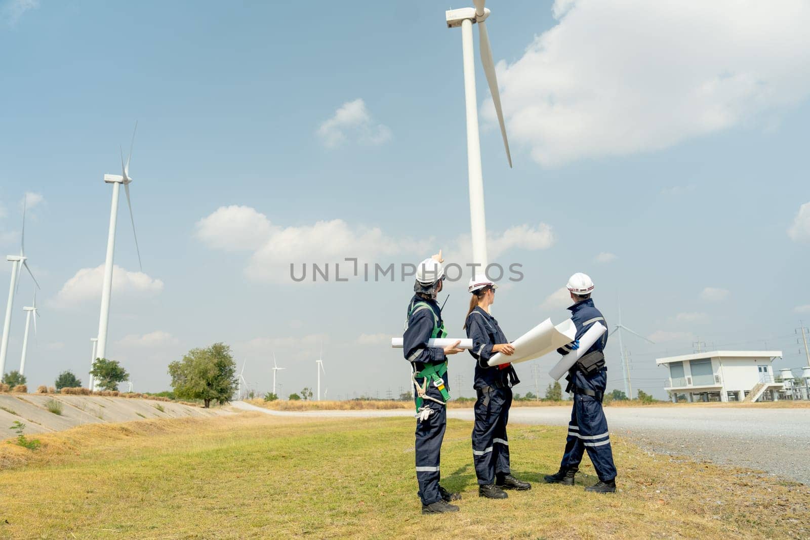 Group of professional technician workers stand with holding project paper plan in front of wind turbine or windmill and look back in area for power plant business.