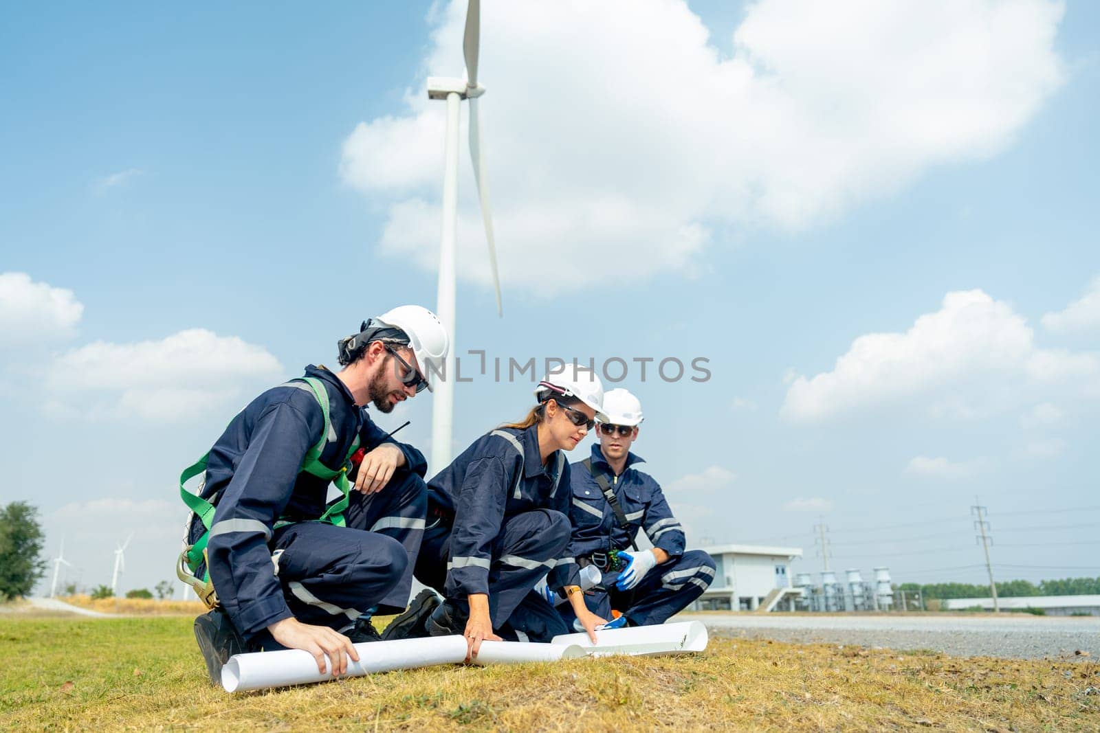 Side view of group professional technician workers sit and discuss work with project paper plan in front of wind turbine or windmill and point to the pole in area for power plant business.