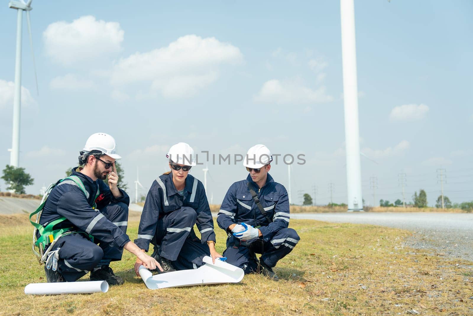 Professional technician workers sit and discuss work with project paper plan in front of wind turbine or windmill and one man point to the paper plan.