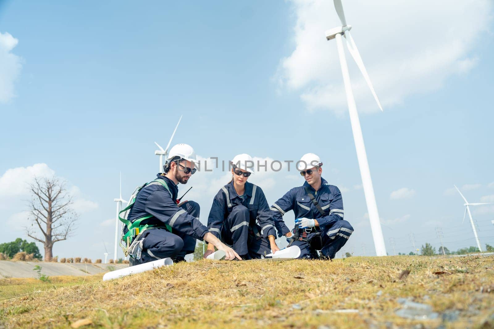 Group of professional technician workers sit and discuss work with project paper plan in front of wind turbine or windmill and point to the pole in area for power plant business. by nrradmin