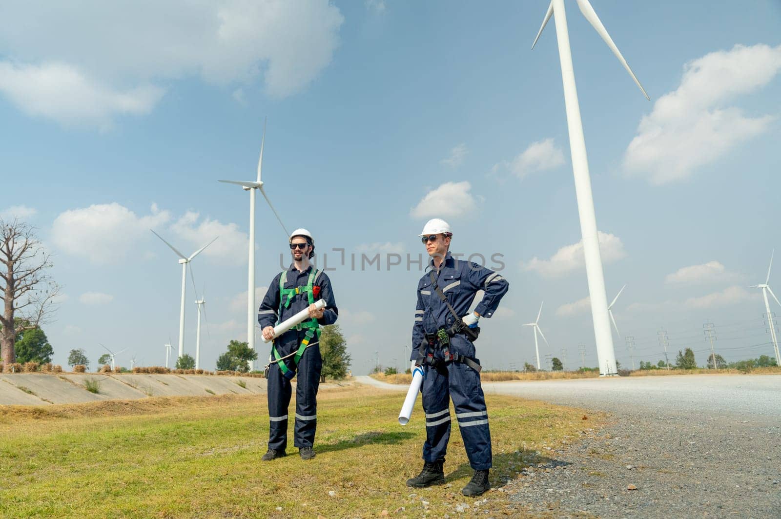 Professional Caucasian workers or technician with safety suit hold construction paper plan and stand in front of wind turbine or windmill in the row as power plant business. by nrradmin