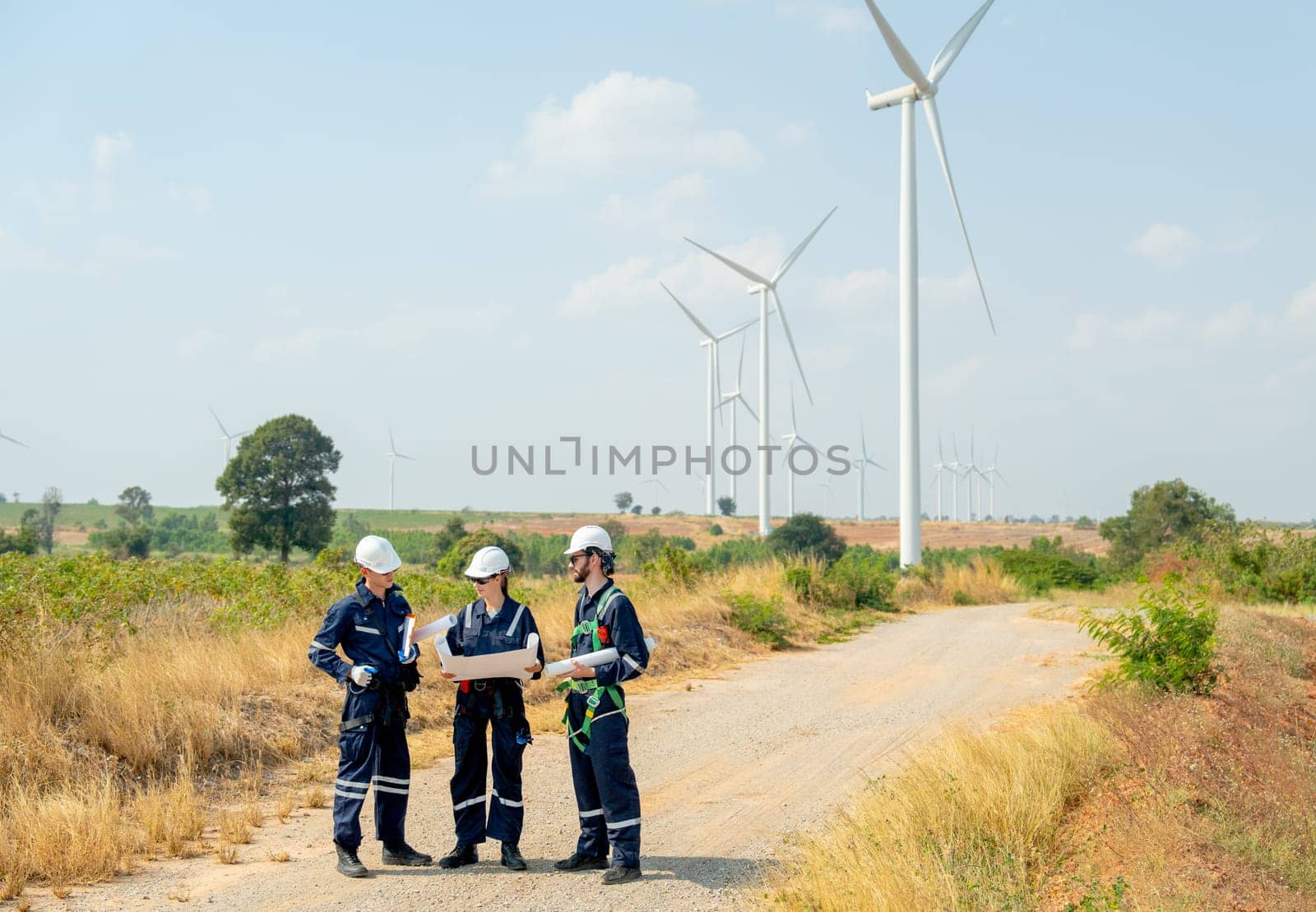 Wide shot group of professional technician workers stand with discuss the project in the paper plan and stand in front of wind turbine or windmill and on road go to maintenance. by nrradmin