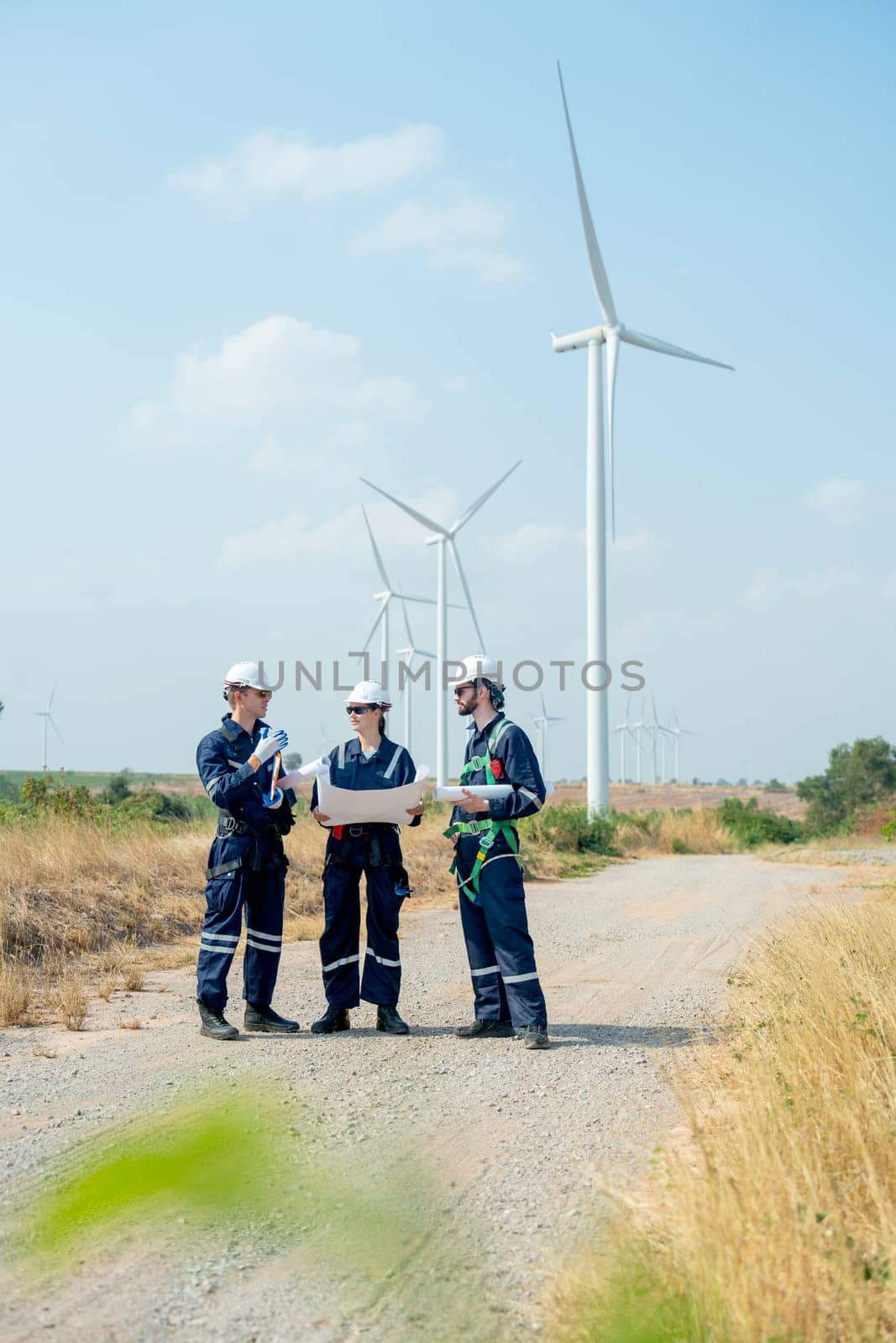 Vertical image of group of professional technician man and woman workers discuss about work project on plan paper in front of wind turbine or windmill in power plant business area. by nrradmin