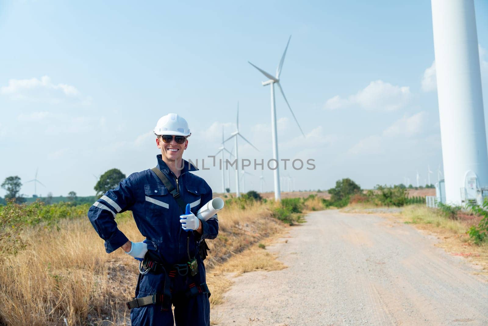 Professional technician worker man with smiling and stand with look at camera in front of wind turbine or windmill with blue sky in concept of green energy for good environment.