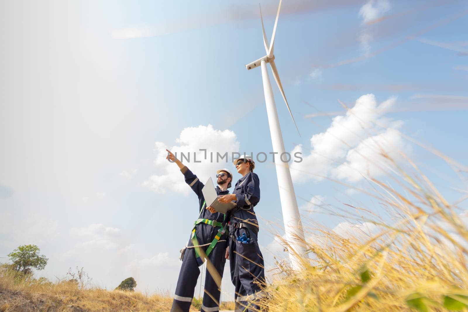 Wide shot professional worker woman hold laptop and stand with her co-worker man point forward and they stand in front of wind turbine or windmill in the field for power plant business. by nrradmin