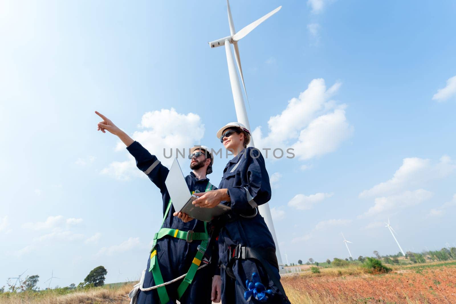 Professional worker woman hold laptop and stand with her co-worker man point forward and they stand in front of wind turbine or windmill in the field for power plant business.