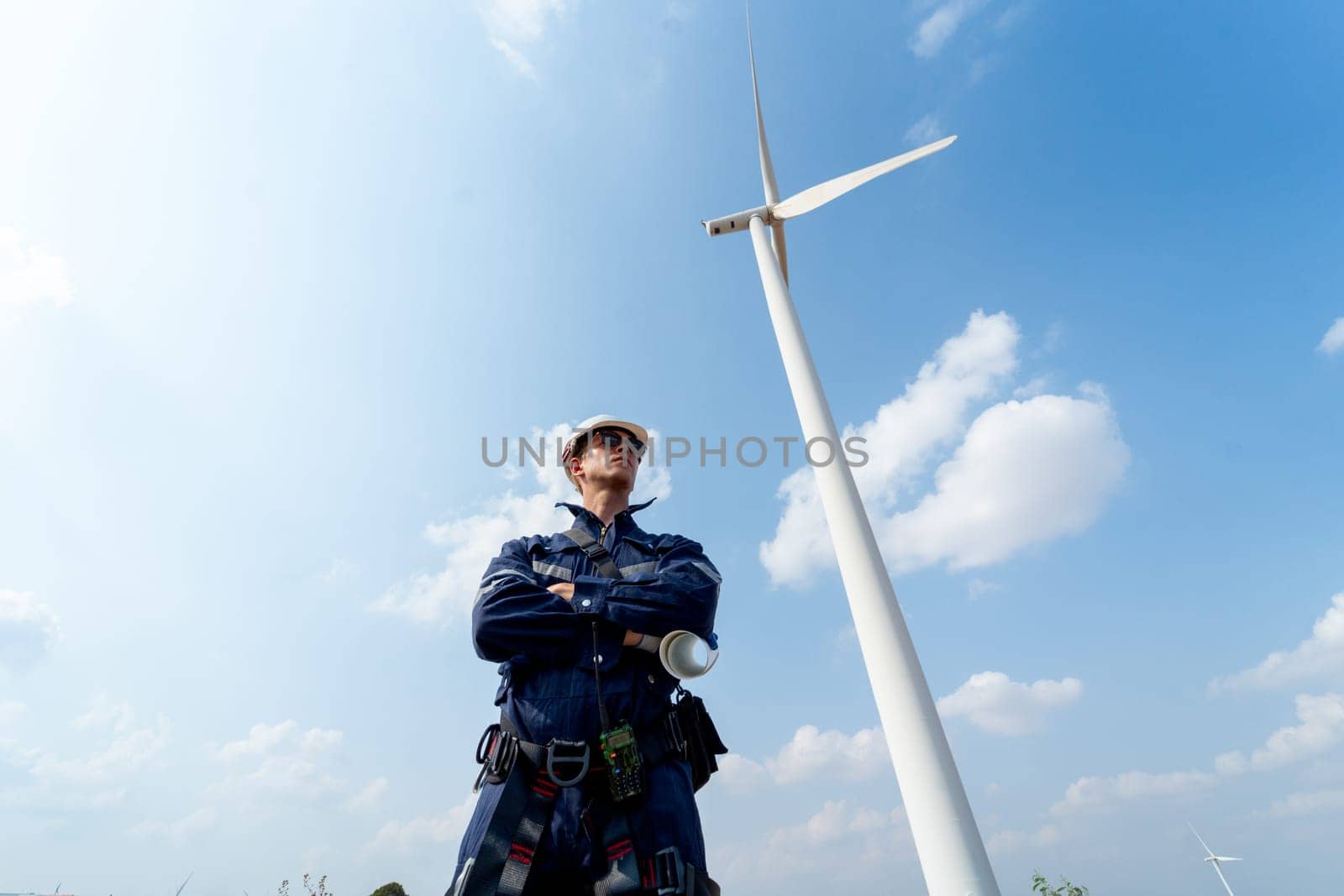 Professional technician worker man stand with arm-crossed and stand with look to right side in front of wind turbine or windmill with blue sky in concept of green energy for good environment. by nrradmin