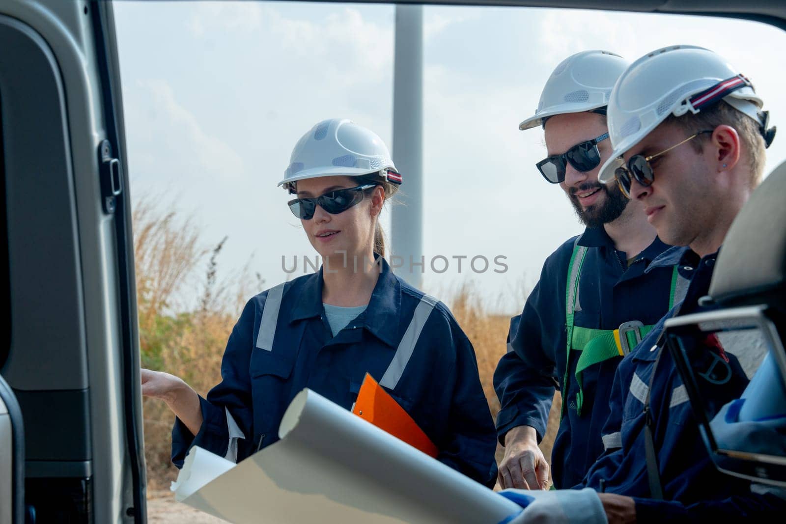 Close up Group of technician workers stand beside the van that open the door and discuss about work using plan paper with windmill or wind turbine on the background.