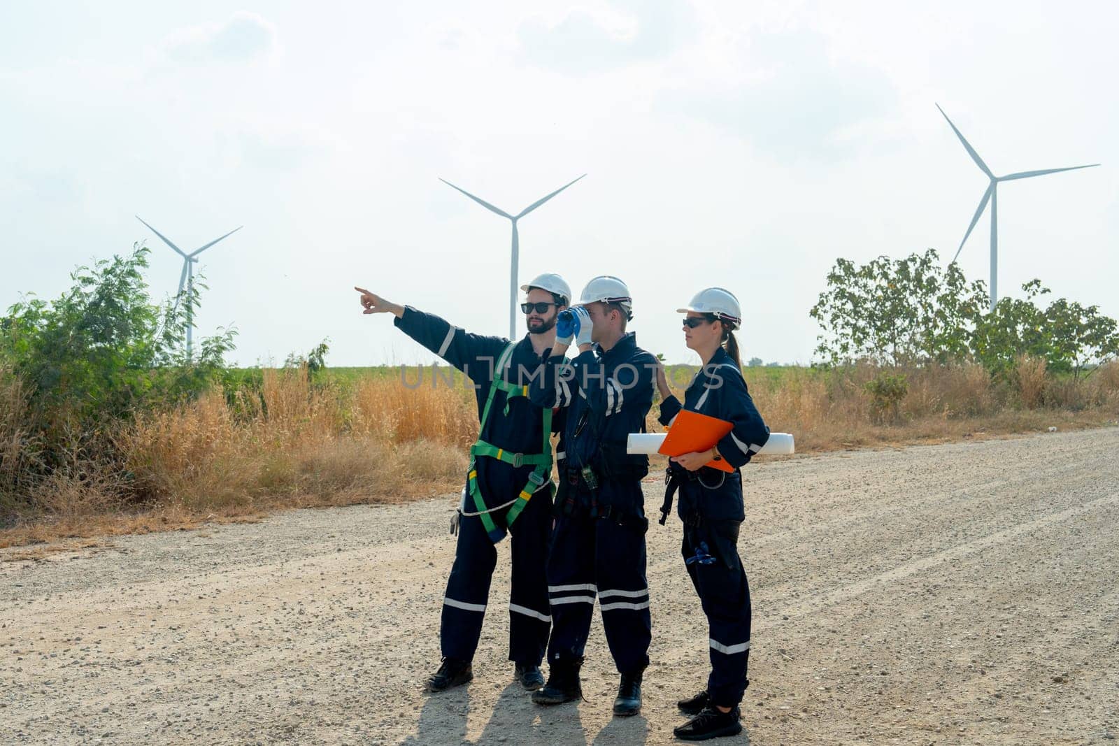 Group of technician workers stand on the road and discuss about work one of them also point to left side with windmill or wind turbine on the background. by nrradmin