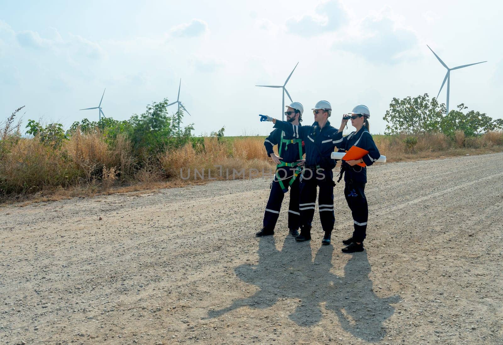 Wide short group of technician workers stand on the road and discuss about work with one of them point to left side and stay with windmill or wind turbine on the background.