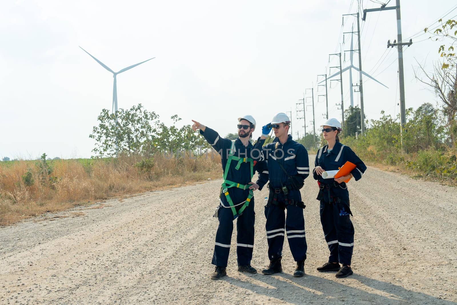 Group of technician workers stand on the road and discuss about work with one of them point to left side and stay with windmill or wind turbine on the background.