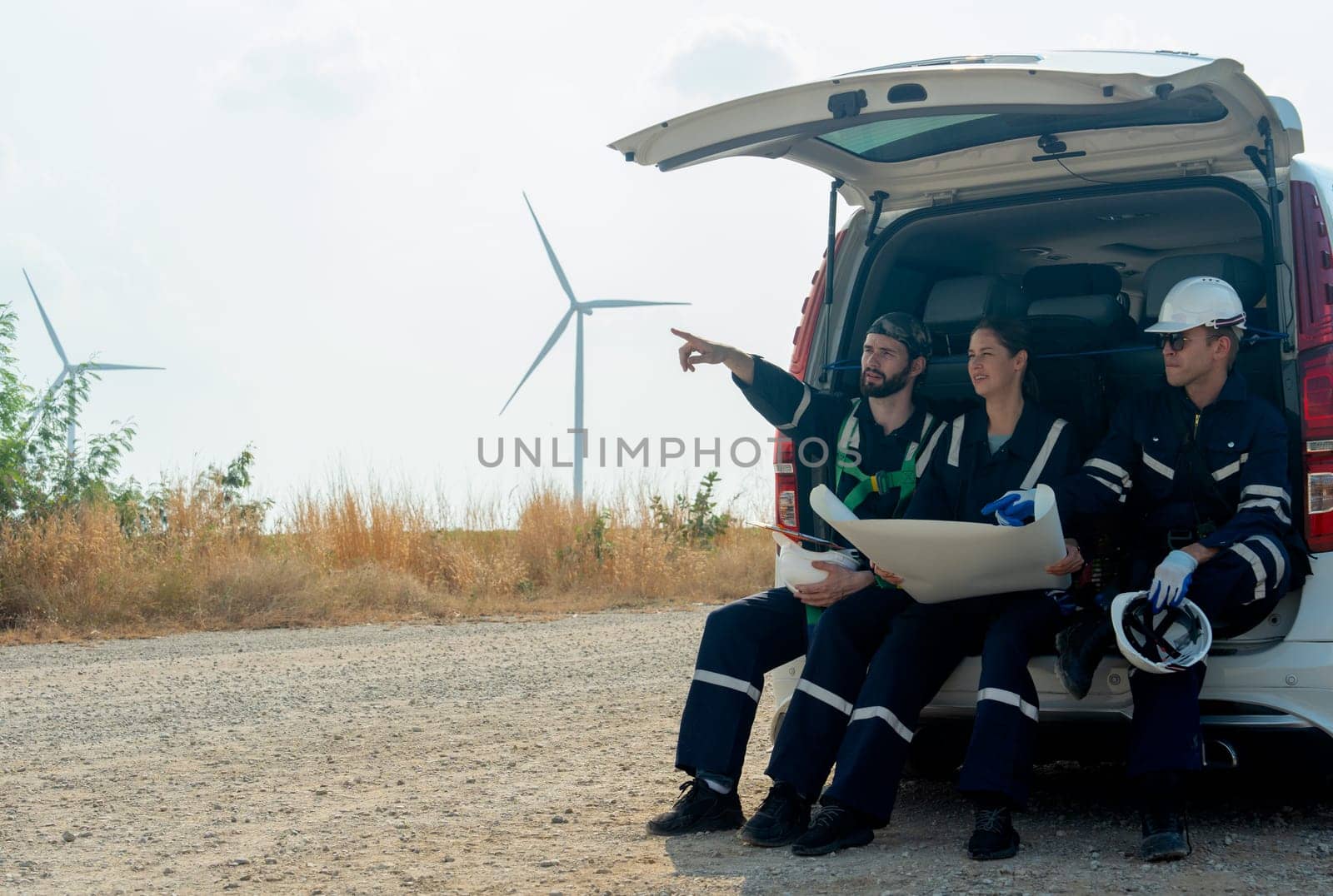 Side view group of technician workers sit on back of van and discuss about work in plan paper on the road with windmill or wind turbine on the background.